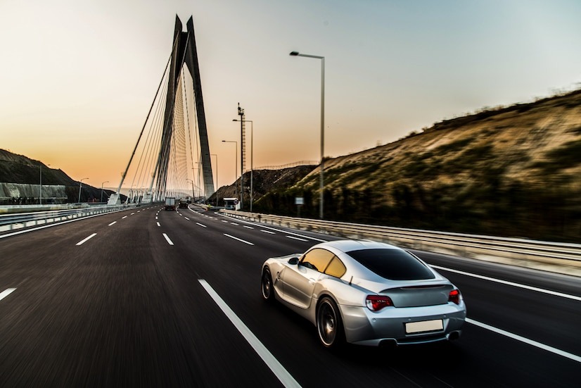 A silver coated car driving on a largely empty highway, dynamic camera perspective, blue and orange hue sky